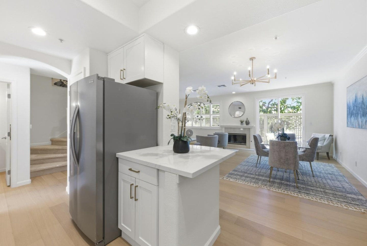 View from the kitchen island across white-oak floors into the dining and living area with a brass chandelier and fireplace