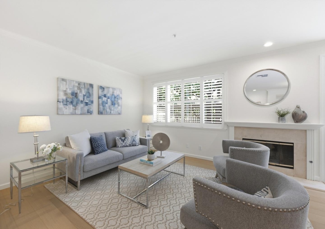 Sunlit living room with a tile fireplace, plantation shutters, and twin upholstered armchairs on a patterned rug