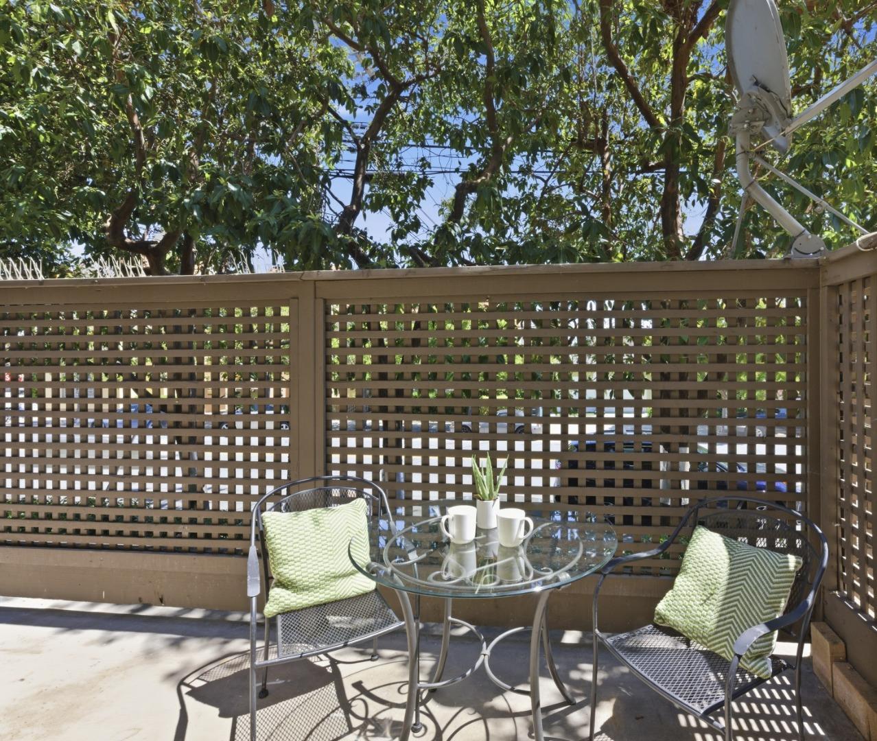 Private patio with a glass bistro table, two chairs, and a lattice privacy screen backed by mature trees